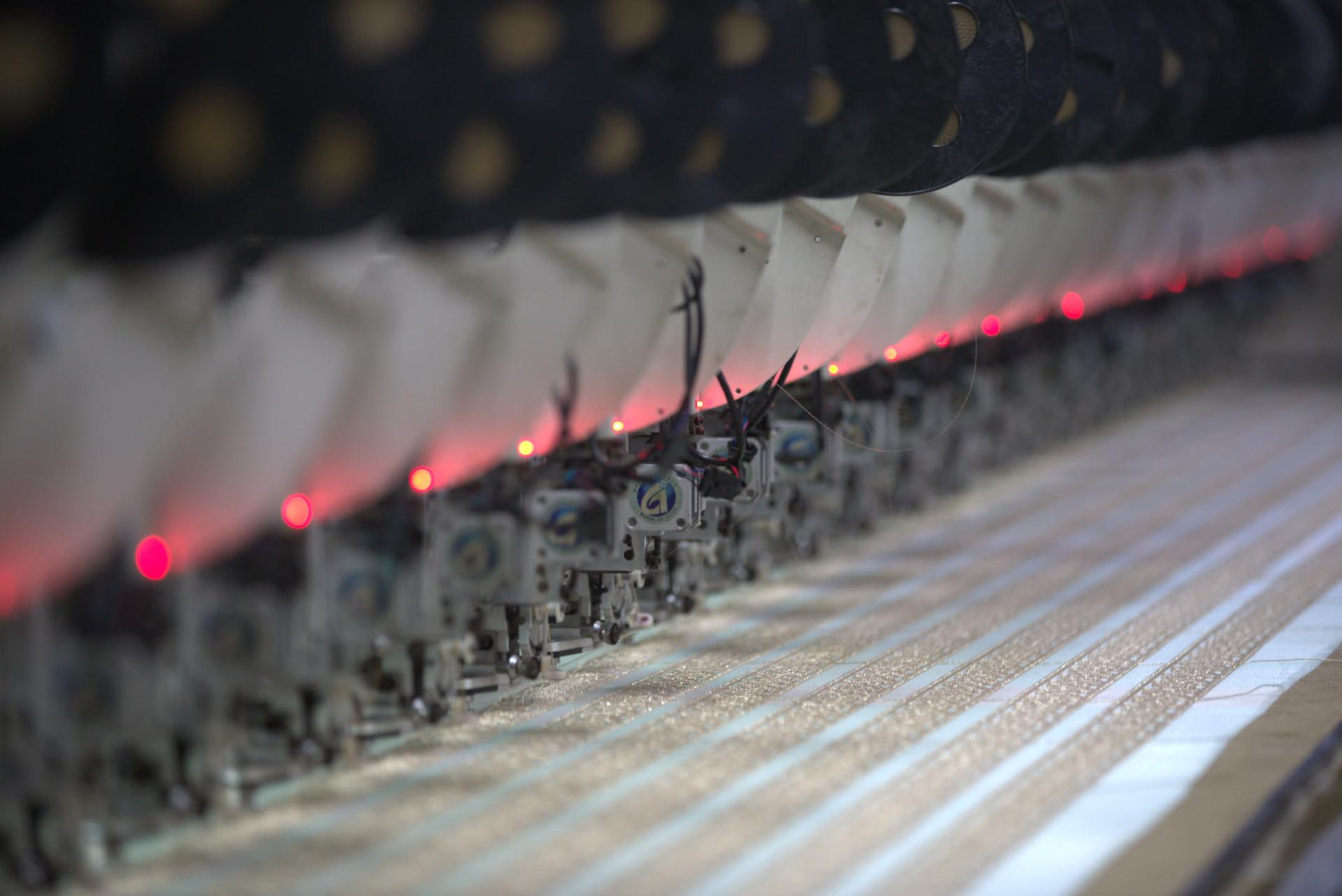 A row of embroidery machine heads mid-run, indicator lamps lit down the line, stitching metallic zari onto lengths of tape.